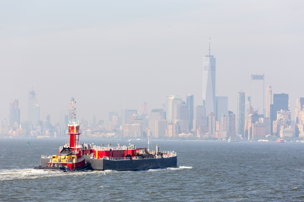 Freight tug pushing cargo ship to the port in New York City and Lower Manhattan skyscarpers skyline in background